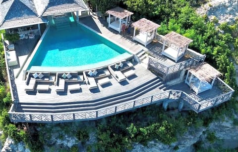 The pool deck from above: a curved pool lined with white loungers, and four thatched cabanas with daybeds